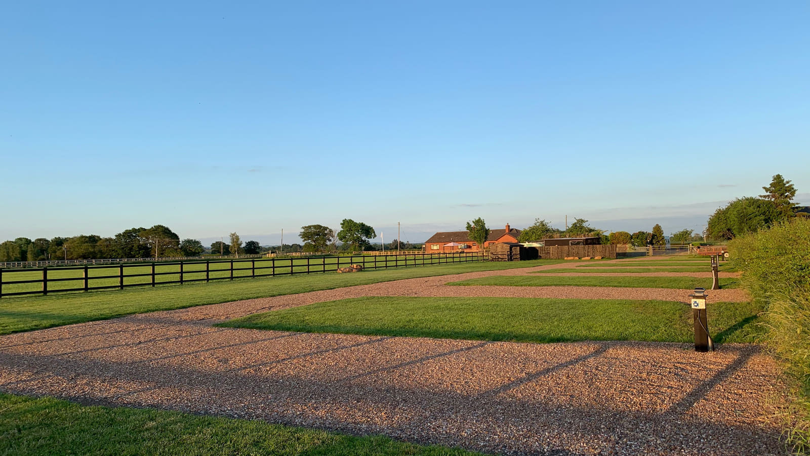 Gravel hard standing pitches with mown grass strips between them at golden hour, black post and rail fencing running out to open pasture, and a timber electric hook up post in the foreground.